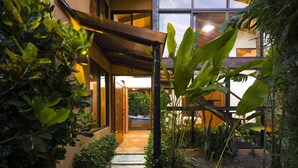 A covered outdoor area with a wooden pergola, surrounded by lush greenery and a view of a modern building with large windows. - Walk to Beach + Shops ~Pool, AC & Concierge (Uvita)