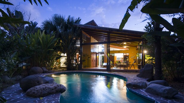 A pool with surrounding rocks and tropical plants, a covered outdoor seating area, and a building with visible interior lighting. - Walk to Beach + Shops ~Pool, AC & Concierge (Uvita)