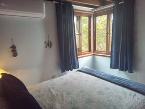 A bedroom with a bed, a window with curtains, a wall-mounted air conditioner, and a wall decoration. - Beach House with Paddle Boards & Water Toys (Maya Beach)