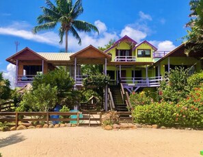 Ein direkt am Strand gelegenes Anwesen mit mehreren Holzhäusern, darunter ein Hauptgebäude mit Balkon und ein kleineres Gebäude mit einer Treppe zu einer Veranda.