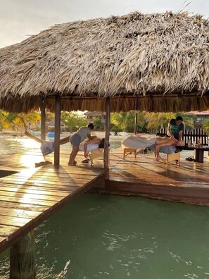 A wooden deck with a thatched roof structure, a wooden bench, and a person lying on a massage table. - Beach House with Paddle Boards & Water Toys (Maya Beach)