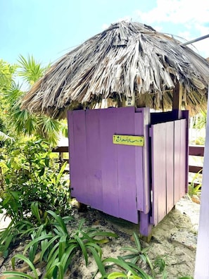 Une petite construction dotée d’un toit de chaume et d’une porte violette, portant l’inscription « Douche », et entourée de verdure.