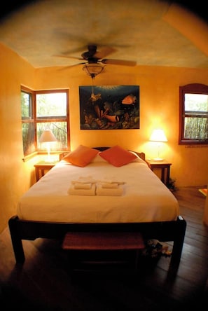 A bedroom with a bed, two bedside lamps, a ceiling fan, and a painting on the wall.