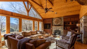Interior acogedor de una cabaña con chimenea de piedra, muebles de madera y ventanas amplias que ofrecen vistas al exterior.