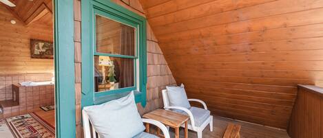 Una terraza de madera con dos sillas blancas y una pequeña mesa, un banco de madera y una ventana con marco verde.