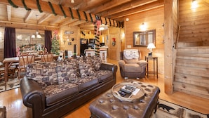 A cozy log cabin interior with a wooden dining area, a sofa with patterned cushions, a leather ottoman, and a kitchen visible through an open doorway. - Spacious 2 BR w/Hot Tub | Pool Table | Wi-Fi (Sevierville)