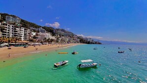 A coastal beach with clear turquoise waters, several boats, and a densely built hillside with multiple buildings.