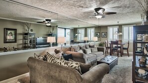 A living room with a large sectional sofa, a coffee table, and a dining area with a table and chairs. There are ceiling fans, a chandelier, and a large window with blinds.