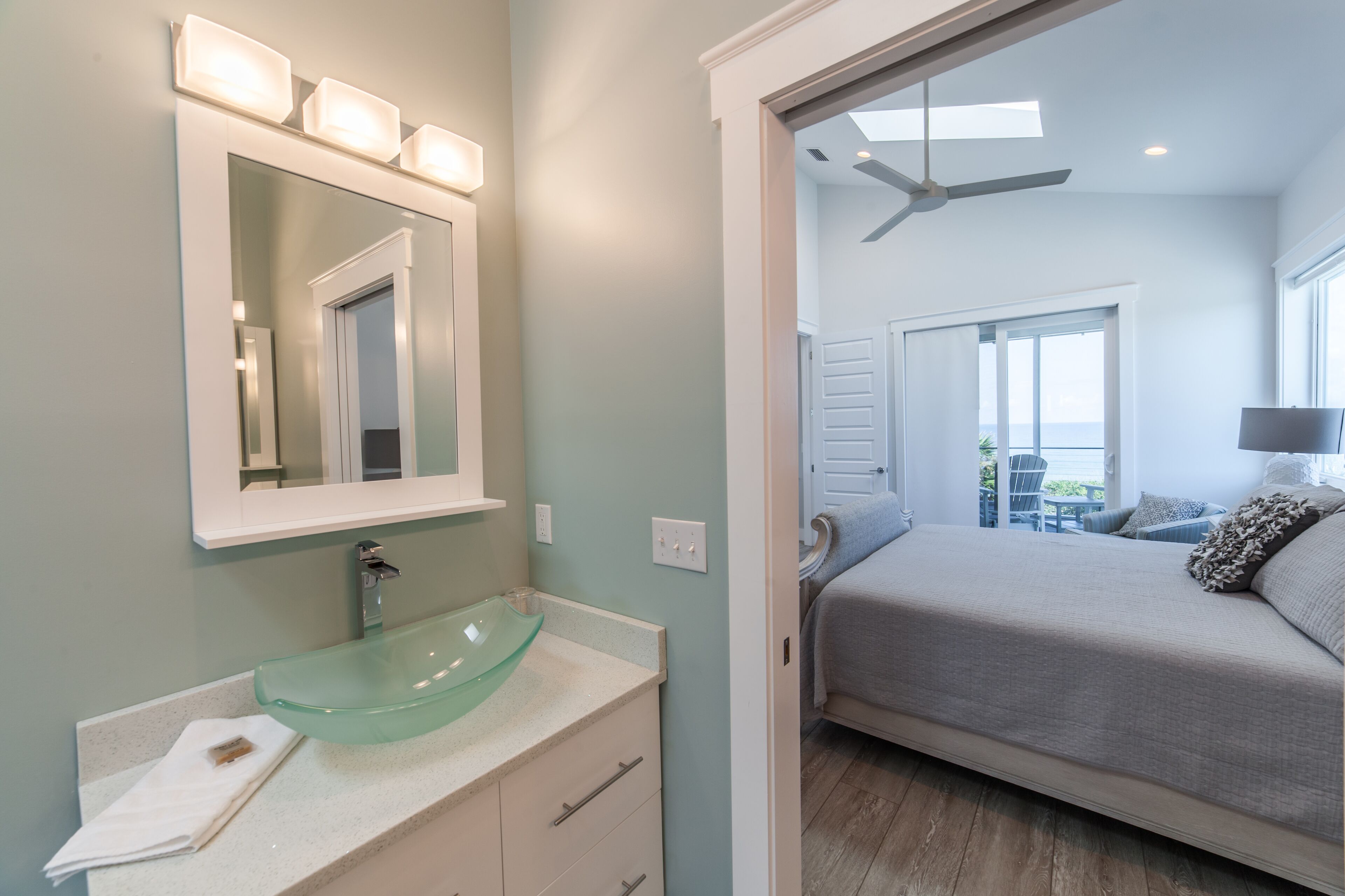 A bathroom with a glass sink, a mirror with lights, and a door leading to a bedroom with a bed and a ceiling fan.