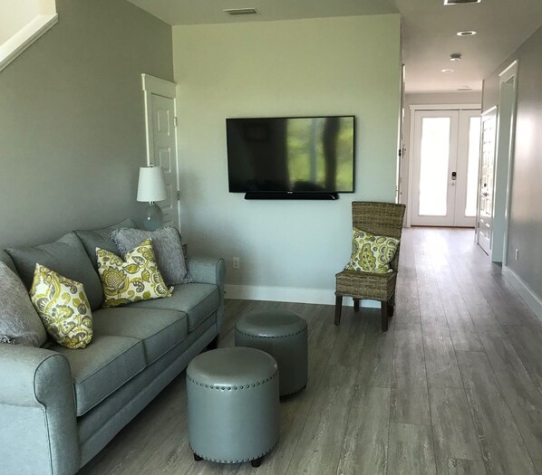 A living room with a grey sofa, ottomans, a wicker chair, and a flat-screen TV mounted on the wall.