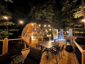 Una terraza de madera con mesa y sillas, rodeada de árboles y una estructura en forma de cúpula geométrica.