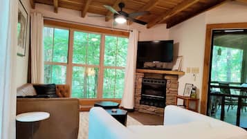 Un salon chaleureux avec une cheminée en pierre, un ventilateur de plafond et une grande fenêtre donnant sur une forêt.