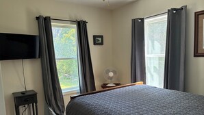 A bedroom with a bed, a TV mounted on the wall, a fan, and two windows with curtains. - Beautiful  camp nestled at the end of a dead end road. (Swanton)