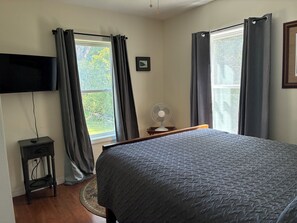 Un dormitorio con una cama, un televisor montado en la pared, un ventilador y dos ventanas con cortinas.