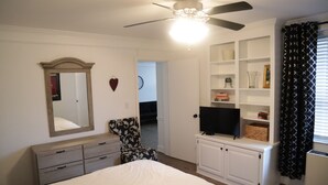Een slaapkamer met een bed, een kaptafel, een dressoir, een stoel, een televisie en ingebouwde planken.