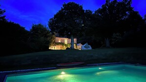 Une piscine avec un bateau, un bâtiment en pierre illuminé, et des arbres en arrière-plan.