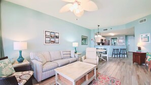 A living room with a sofa, coffee table, and chairs, a dining area with a table and chairs, and a kitchen with a refrigerator and stove. - Beach Club Ground Floor! Right on the beach - Pools- WIFI (Gulf Shores)
