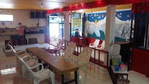 A dining area with a long wooden table and plastic chairs, a small stage with a speaker, and a mural depicting a mountain scene. - Om Ganesh Hostel - Adults Only (Ko Pha-ngan)