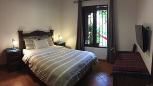 A neatly made bed with striped bedding, two bedside lamps, a window with curtains, and a small bench with a colorful mat. - Apartamentos los Nazarenos PRIVATE APT 1 - heated swimming pool, jacuzzi and gym (Antigua Guatemala)