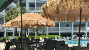 A poolside area with lounge chairs and thatched umbrellas, a pool, and a building with balconies in the background. - Happy Place Gated ResortBldg.1,First floor, pool, deli/gift shop, bar (Hilton Head)