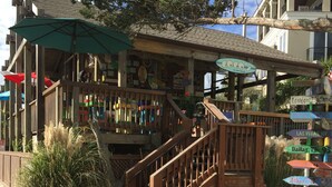 A wooden building with a staircase leading to a deck, surrounded by sand and plants. - Happy Place Gated ResortBldg.1,First floor, pool, deli/gift shop, bar (Hilton Head)