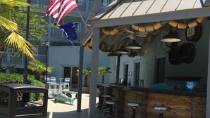 A beachside bar with a rustic wooden counter and stools, an American flag, and a view of a multi-story building in the background. - Happy Place Gated ResortBldg.1,First floor, pool, deli/gift shop, bar (Hilton Head)