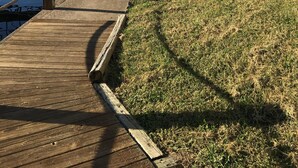 Une promenade en bois menant à un quai, avec une berge herbeuse et un bâtiment doté de balcons.