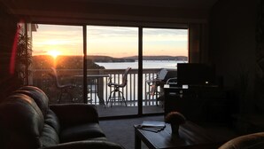 Un salon avec un canapé, une table basse et un téléviseur. Il y a un balcon d’où l’on peut admirer le coucher de soleil sur une étendue d’eau.