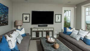 Ein modernes Wohnzimmer mit einem großen Flachbildfernseher, grauen Sofas mit blauen Kissen, einem Couchtisch mit Dekoration und einem Fenster mit Blick ins Freie.