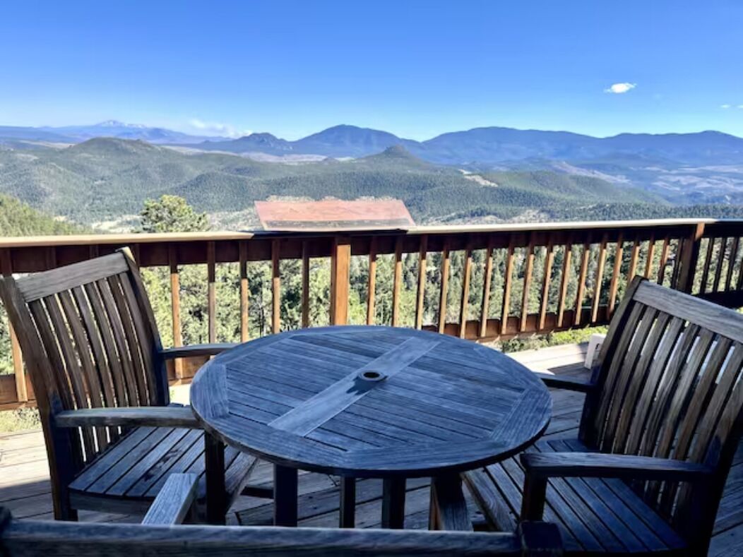 Eine Holzterrasse mit einem runden Tisch und zwei Stühlen, mit Blick auf eine gebirgige Landschaft.