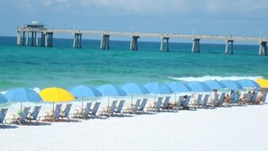 A beach with rows of sun loungers and umbrellas, a pier extending into the ocean, and a small structure on the pier. - Destin West Beach Resort Villa Studio (Fort Walton Beach)