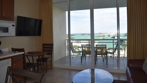 A hotel room with a kitchenette, a flat-screen TV, a dining area with chairs and a table, and a balcony with a view of buildings. - Destin West Beach Resort Villa Studio (Fort Walton Beach)