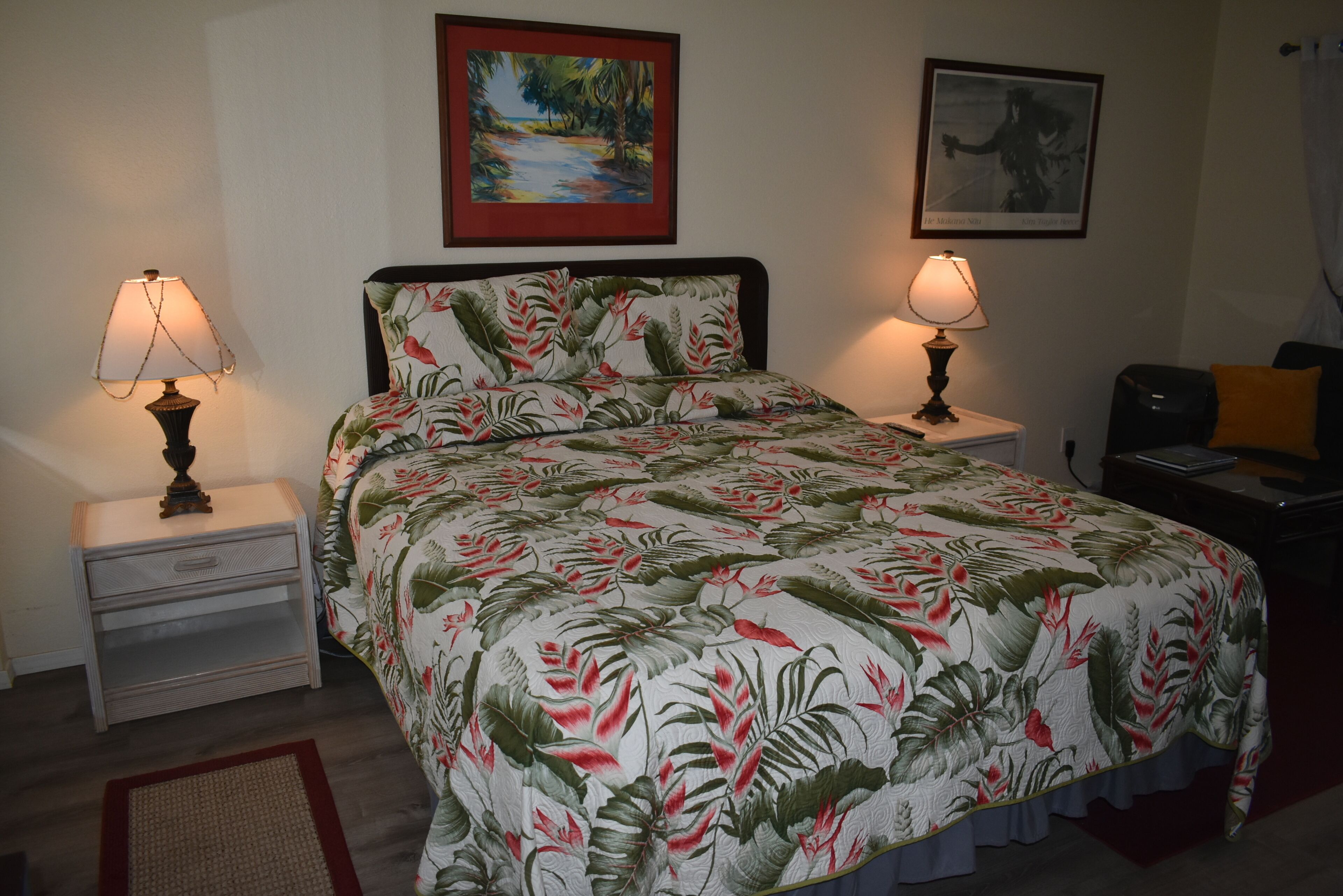 Una cama bien hecha con ropa de cama floral, dos mesitas de noche con lámparas, un cuadro enmarcado sobre la cama y un altavoz pequeño a la derecha.