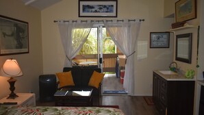 A compact hotel room with a bed, a small table, a sofa, a desk, a kitchenette, and a balcony with a view of palm trees. -  Fresh & Clean, Air Conditioned Second Floor Condo in Princeville  (Princeville)