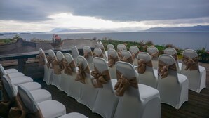 Rows of white chairs with brown bows, arranged on a wooden deck with a view of a large body of water and distant mountains.