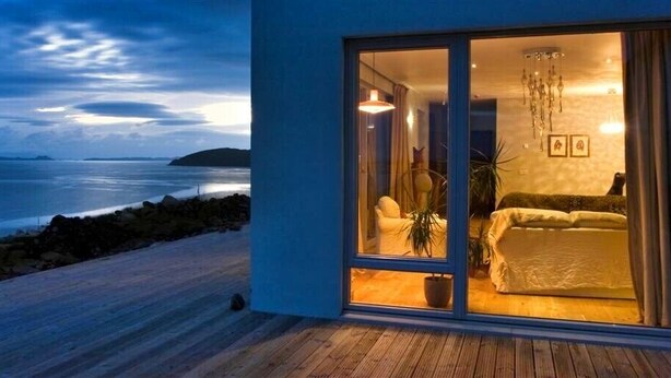 A modern bedroom with a large sliding glass door leading to a balcony overlooking the sea.