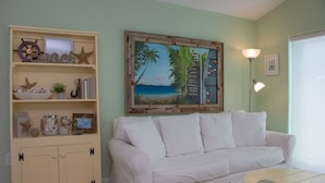A room with a white sofa, a wooden cabinet with decorative items, a framed beach painting, and a floor lamp. - Flamingo Nook. Renovated, Private Pool, Canal & Cabana Club Access! (Key Colony Beach)