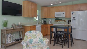 A kitchen with wooden cabinets, a microwave, a refrigerator, a dining table with stools, and a television mounted on the wall. - Flamingo Nook. Renovated, Private Pool, Canal & Cabana Club Access! (Key Colony Beach)