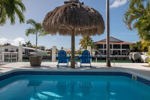 A swimming pool with blue tiles, two blue chairs, a thatched umbrella, and a building with a red roof in the background. - Flamingo Nook. Renovated, Private Pool, Canal & Cabana Club Access! (Key Colony Beach)