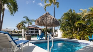 A swimming pool with lounge chairs, a thatched umbrella, and palm trees. - Flamingo Nook. Renovated, Private Pool, Canal & Cabana Club Access! (Key Colony Beach)