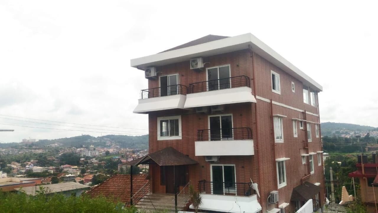Un immeuble à plusieurs étages, doté d’un toit plat, de balcons à chaque étage et offrant une vue sur un paysage vallonné en arrière-plan.