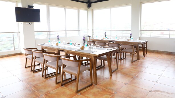 A spacious room with large windows, a flat-screen TV mounted on the wall, and multiple tables with chairs. - Lali Petals (Madikeri)