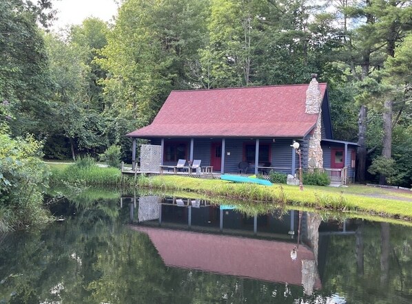 A cabin with a red roof, a porch, and a small dock extending into a pond.