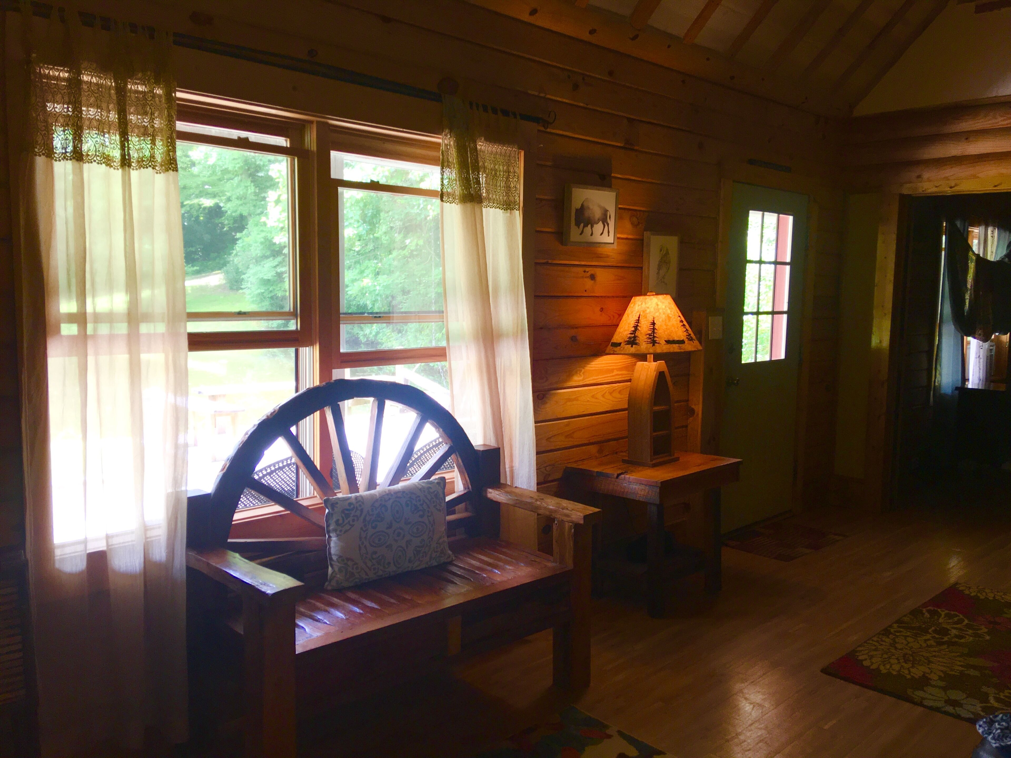 Un interior de cabaña acogedor con un banco de madera, una mesa con una lámpara y una ventana con cortinas.