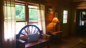 Un interior de cabaña acogedor con un banco de madera, una mesa con una lámpara y una ventana con cortinas.