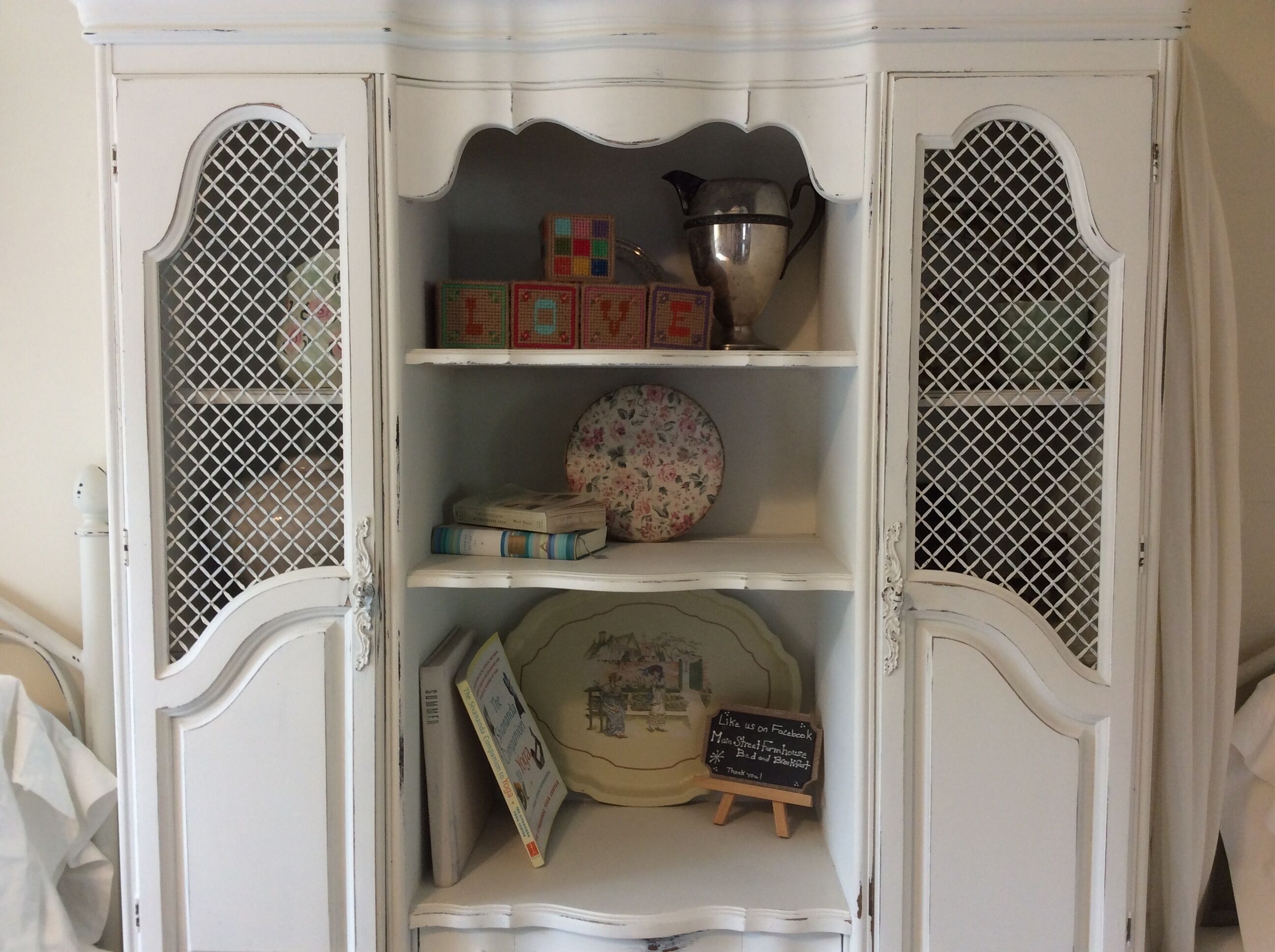 Ein weißer Schrank mit verzierten Metalldetails, Regale mit antiken Gegenständen und ein Kreidetafel-Schild.