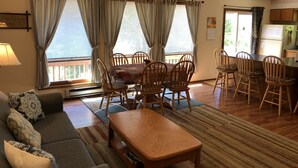 Sala de estar com sofá, mesa de jantar de madeira, cadeiras e área de cozinha visível pela janela.