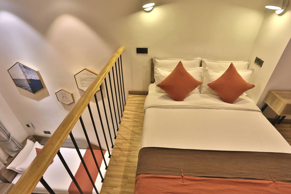 Habitación de hotel con una escalera de madera, una cama con ropa de cama blanca y naranja, y una pared decorada con cuadros.