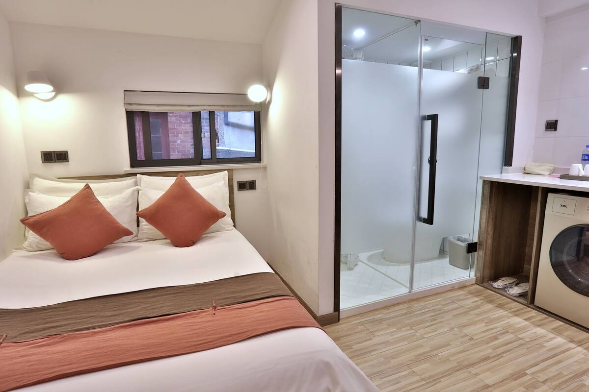 Habitación de hotel con cama, ventana, cabina de ducha de vidrio y lavadora.
