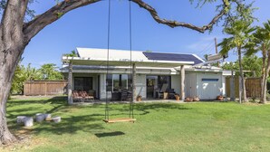 Une maison avec un grand arbre au premier plan, une balançoire et un espace patio aménagé avec du mobilier d’extérieur.